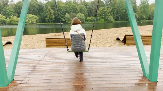 A girl sits on a swing overlooking a calm lake in Pushkin, Saint Petersburg. She feels peaceful and relaxed in nature. The quiet scene brings serene comfort.