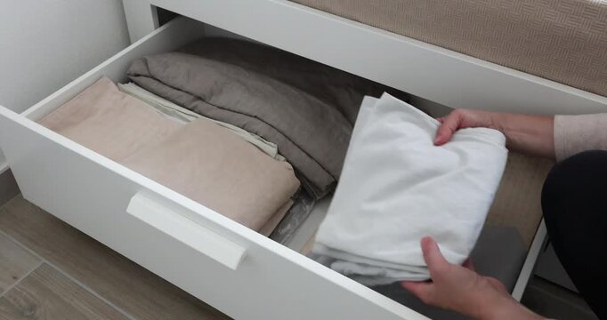 Woman neatly organizes folded bedsheets inside underbed drawer. Minimalist housekeeping routine brings tidy aesthetic comfort indoors. Smart home organization hacks optimize bedroom spaces efficiently