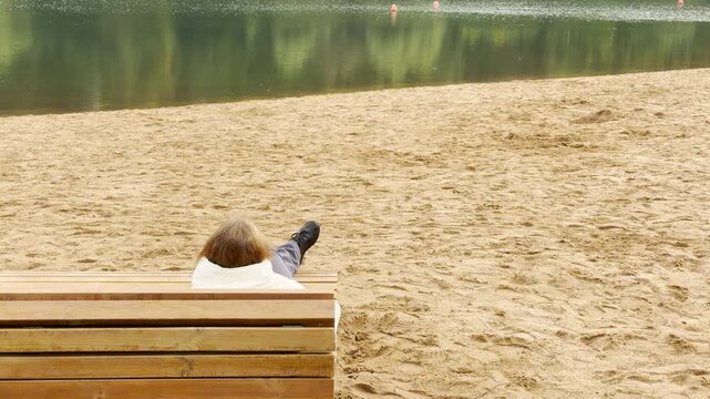 A woman relaxes on a wooden bench along the sandy shore in Pushkin, Saint Petersburg. She gazes out toward the tranquil water. Her quiet rest brings a calm, peaceful feeling.