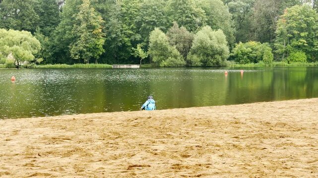 A young child plays by the water edge on a sandy beach in Pushkin, Saint Petersburg. He feels peaceful joy near the quiet pond. Green trees offer calm comfort.