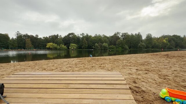 A child walks along the sandy shore by a quiet lake in Pushkin, Saint Petersburg. Toy trucks rest on the beach under a soft overcast sky. The tranquil view brings a peaceful and calm feeling.