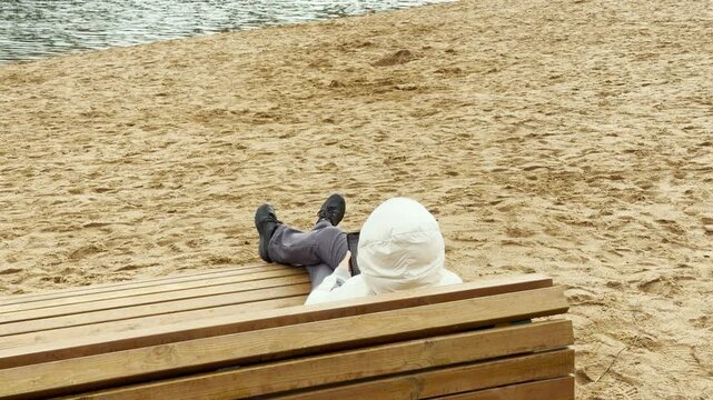 A person rests comfortably on a wooden lounger in Pushkin, Saint Petersburg. They relax peacefully on the sandy beach. The quiet environment brings serene comfort.