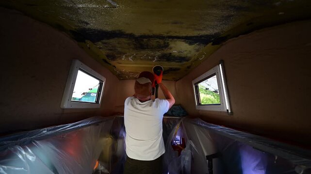 Man with a respirator mask sanding the fiberglass ceiling of a camper van