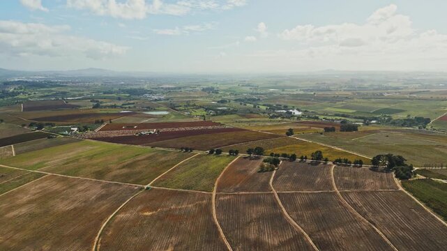Aerial view of cultivated farmland clearly displays colorful fields, parallel tillage lines, rural roads, scattered trees, small buildings, and distant low hills beneath partly cloudy skies.