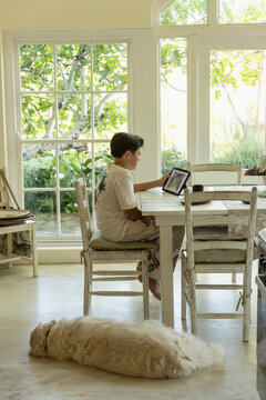 13 year old boy sitting at table in light filled room, using tablet computer early morning