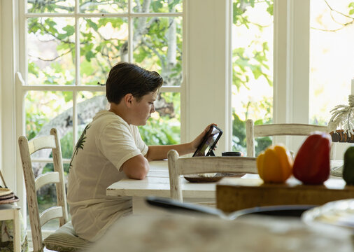 13 year old boy sitting at table in light filled room, using tablet computer early morning
