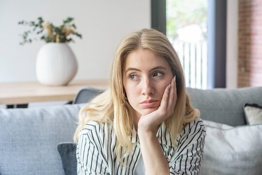 Young woman sitting on a gray couch in a living room, propping her head with her hand, looking away with a pensive and tired expression, conveying feelings of sadness and boredom