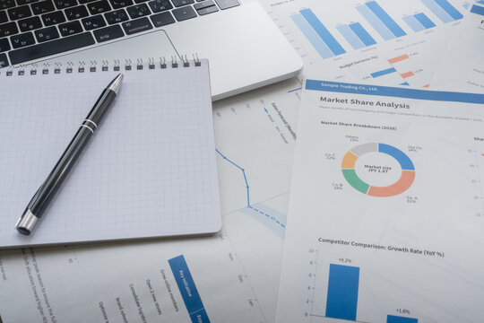 Blank grid notebook and pen on printed market share charts beside a laptop, with copy space.