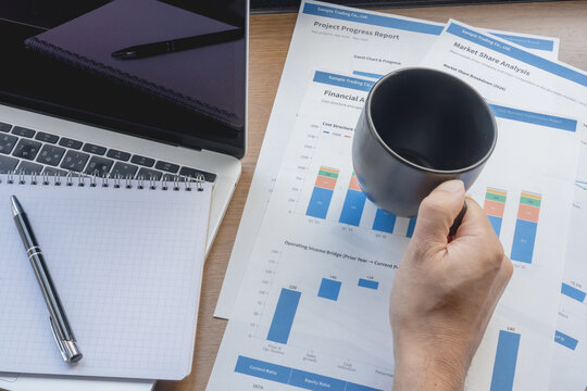 Hand holding a black mug over financial reports beside a blank grid notebook, pen and laptop.
