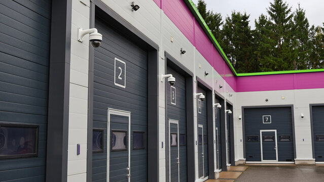 Modern self storage facility featuring a row of numbered gray garage doors with integrated pedestrian doors, each unit protected by a mounted surveillance camera, under a pink and green roof line