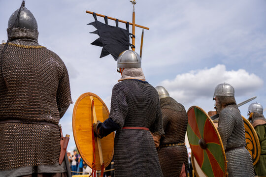 Medieval reenactors in chain mail and helmets stand with wooden shields beneath a black banner at an outdoor historical display.