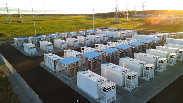 Large battery energy storage park with rows of containerized BESS units and nearby electrical substation at golden sunrise, supporting renewable grid stabilization and decarbonization