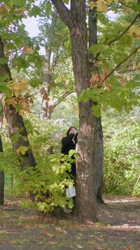 Woman walking among trees in autumnal woodland setting. Woman holds object while moving between trunks under dappled sunlight with yellow and green foliage