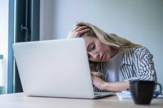 Exhausted young woman leaning on her desk with headache and fatigue, struggling to focus on laptop during long workday at a home office, overwhelmed and overworked