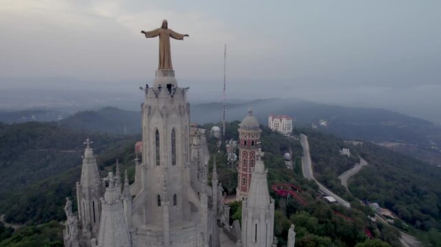 4K aerial drone footage of the Temple of the Sacred Heart on Tibidabo, Barcelona, featuring the iconic Christ statue, historic basilica and lush green mountain landscape at sunset.