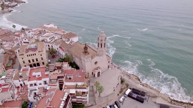 4K drone aerial view of Sitges, Spain, featuring the iconic Sant Bartomeu i Santa Tecla church overlooking the Mediterranean coast, beaches and historic seaside town.