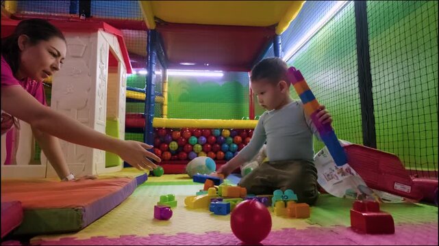 Dolly in shot of a two-year-old boy stacking vibrant building blocks while sitting on soft mats at an indoor playground with his mother in Mexico City.