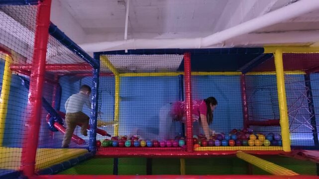A two-year-old Latino boy and his mother play inside a colorful indoor playground, navigating a ninja obstacle course with ball pits and ropes in Mexico City.