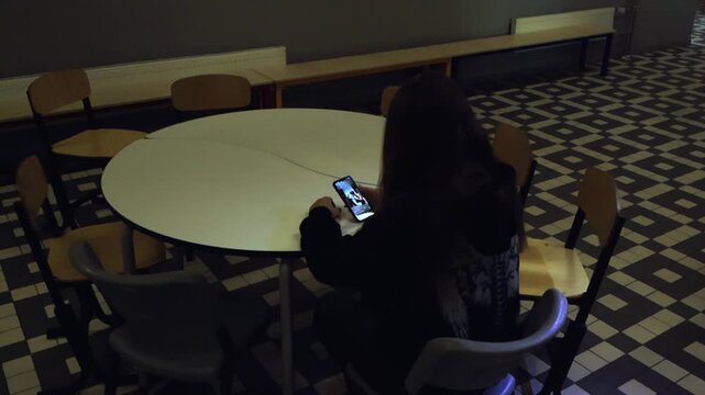 Solitary young woman sitting alone at a round table in an empty, dimly lit school hallway, endlessly scrolling through her social media feed on a smartphone, ignoring her surroundings