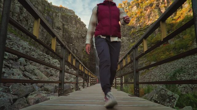 Female adventure traveler walking toward the camera on a wooden boardwalk with spectacular Njupesk&auml;r Waterfall in the background, surrounded by vibrant autumn colors in Fulufj&auml;llet.
