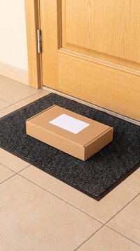 Cardboard parcel with blank white shipping label on black doormat in front of wooden door, home delivery, online shopping, courier package and e-commerce order arrival concept