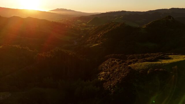 Aerial establishing view of Raglan, New Zealand, at sunset, showing rolling hills, coastal landscape, distant mountains, and warm golden evening light.