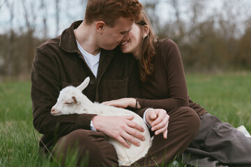 Naklejka premium A couple sits in a grassy field outdoors, warmly embracing a small goat kid resting on their laps, with tender contact and bright smiles conveying simple lifestyle joys and animal companionship.