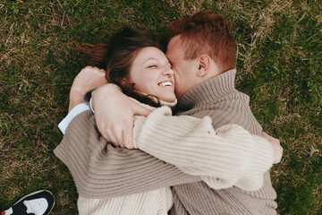 Naklejka premium Romantic couple lying on grass, embracing and kissing, wearing knit sweaters, soft natural light, candid outdoor moment, warm cozy mood, lifestyle scene of affection and togetherness