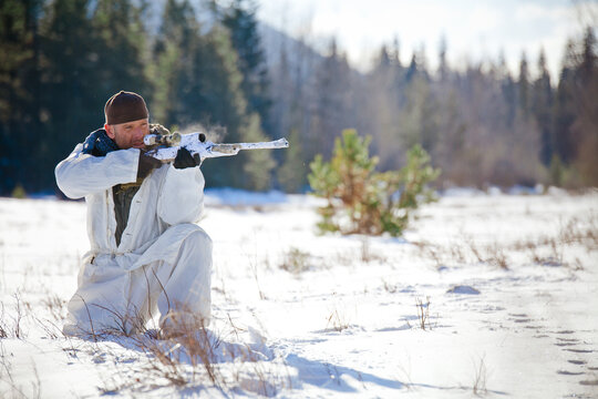 Man in white winter camouflage aims sniper rifle in snow
