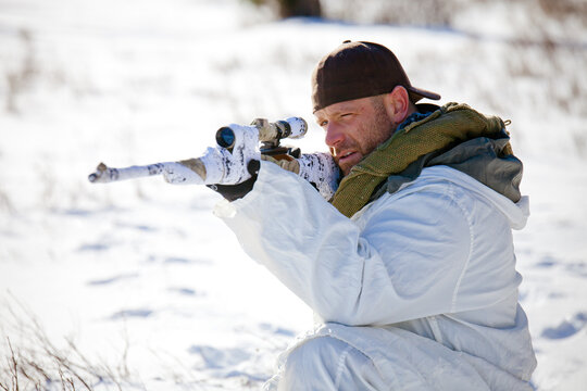 Man in white winter camouflage aims a sniper rifle in snow