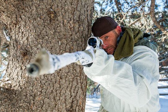 Winter sniper aims his rifle from behind a tree