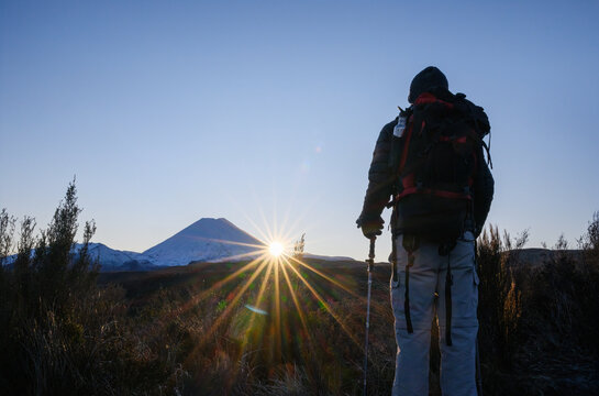 Hiker looking at Mount Ngauruhoe as sun starbursts shining through at sunrise. Taranaki Falls Track in winter. Tongariro National Park. New Zealand.