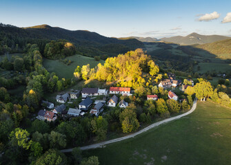 Fototapeta premium Drone view of small mountain village in Slovakia surrounded by forests, and rolling hills in vibrant fall colors, bathed in golden afternoon light.