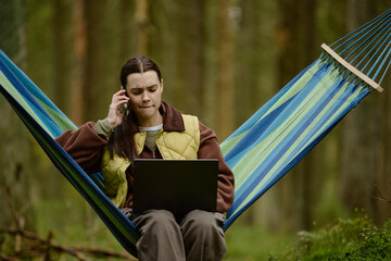 Young adult Caucasian woman sitting in hammock in forest, talking on smartphone while working on...