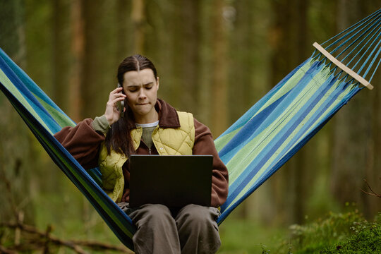 Young adult Caucasian woman sitting in hammock in forest, talking on smartphone while working on laptop. Visual representing remote work stress suitable for telecommuting campaigns