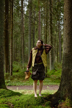 Vertical image of young adult Caucasian woman standing barefoot on moss in forest holding boots. Image suitable for outdoor wellness, ecotourism and mindfulness campaigns