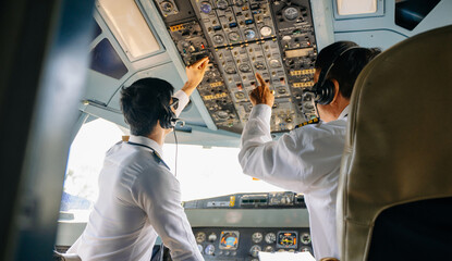 Two airline pilots adjust switches on the overhead control panel inside an airplane cockpit during a preflight check. © Nuttapong punna