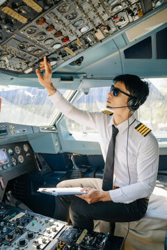 A focused airline pilot wearing a headset checks instrument gauges on the cockpit dashboard with a clipboard during preflight.