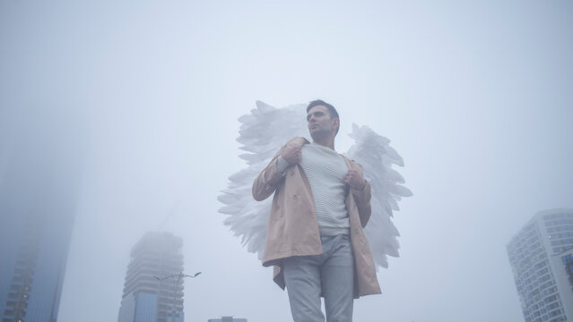 A low angle conceptual shot of a mid adult man wearing large white angel wings and beige coat posing against blurred modern skyscrapers hidden in dense fog, urban lifestyle background