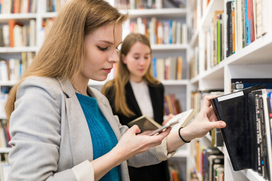 Portrait of teenage girl looking at cell phone while taking book of shelf in a public library