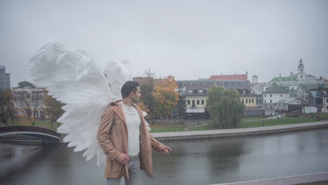 Side view of man with white angel wings looking at autumn city riverfront with copy space
