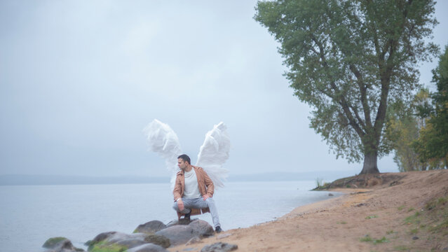 Man with white angel wings crouching on rocks by misty lakeside shore with copy space