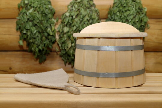 On a plank bench, next to a sauna hat, stands a wooden barrel with cover, against the background of log wall with a birch brooms.
