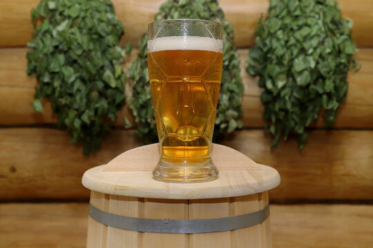 On a top of a wooden barrel stands a glass goblet of light beer, against the background of a log wall with a birch brooms.