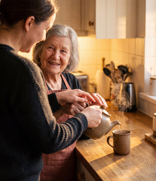 Caring adult daughter helping her happy elderly mother pour hot tea from a ceramic teapot in a warm kitchen