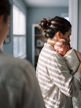 A tender moment of a mother holding her sleeping newborn baby wrapped in a blanket in a sunlit living room at home