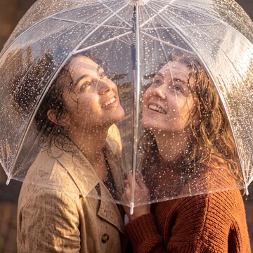 Two cheerful young women looking up together under a clear umbrella with raindrops during a sunny autumn rain shower