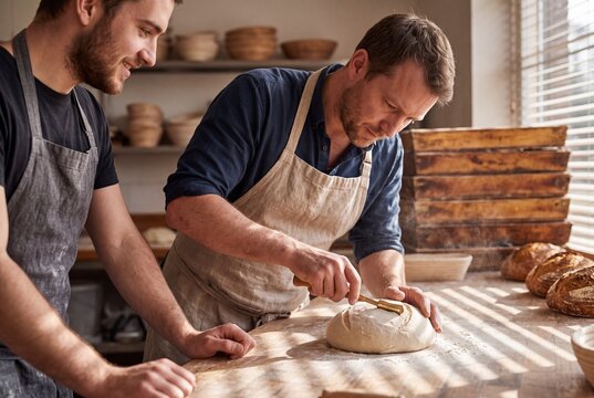 Two male artisan bakers scoring a loaf of sourdough dough with a lame knife in a craft bakery workshop