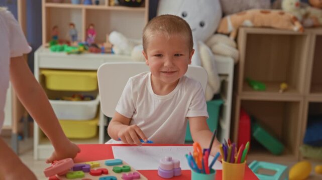 Brother and sister drawing with crayons at a red table, boy holds paper up to show scribbles in a playroom inside a building; sibling bonding joy.