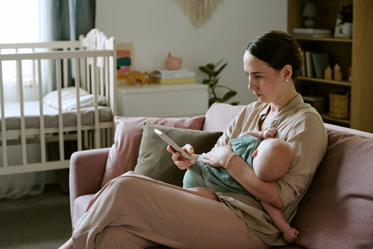 Caucasian young adult mother holding baby child while using smartphone sitting on couch at home. Image suited for parenting mobile application promotion and telehealth services advertising